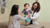 Female physician examines toddler while mother holds him in standing position.