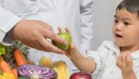 Little boy takes apple from doctor's hand, next to table with vegetables.