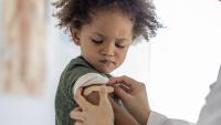 A young boy of African decent sits up on an exam table as his female doctor gives him a vaccination.